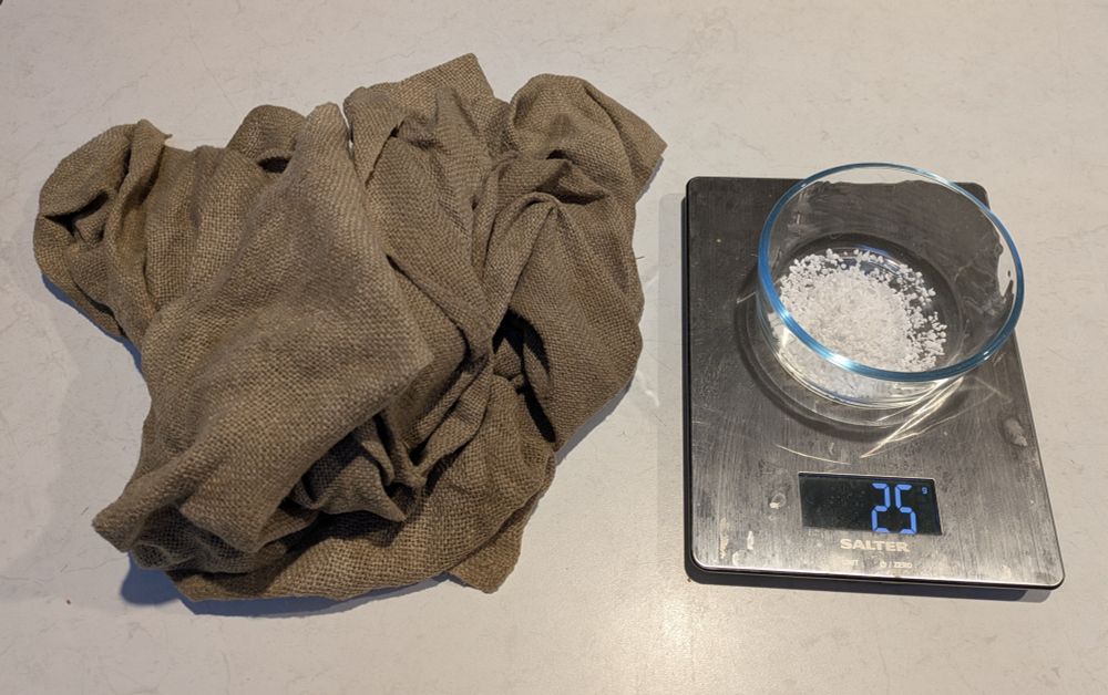 A piece of tannin-stained brown linen sits next to a scale. On the scale is a small glass bowl containing 25g of aluminium sulphate intended as a mordant for the linen.