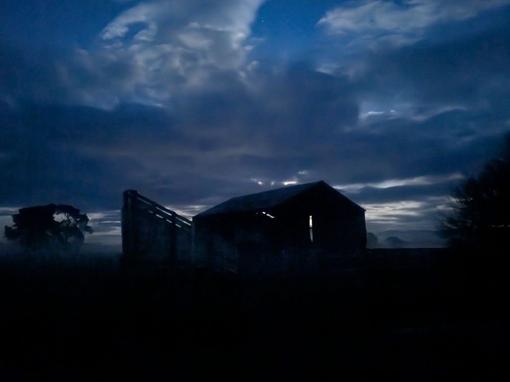 An old barn and cattle loading ramp, silhouetted against a cloudy sky with the light of the full moon backlighting it all