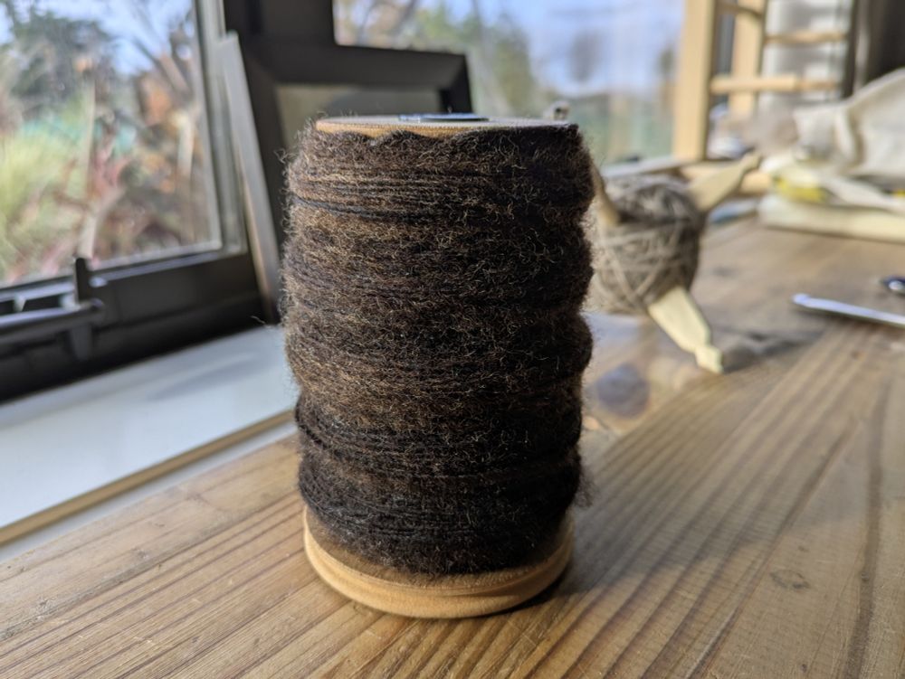 A spool of very dark brown single yarn about 1mm diameter sits on a wooden shelf with a turkish spindle in the background. The wool is spun semi-worsted and has a slight golden halo.