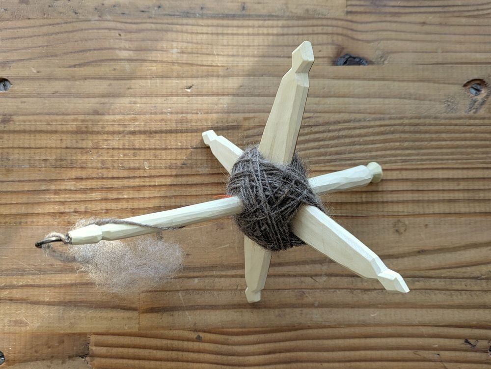 A small Turkish drop spindle made of pale hazel wood, with some medium grey corriedale single wound onto it.