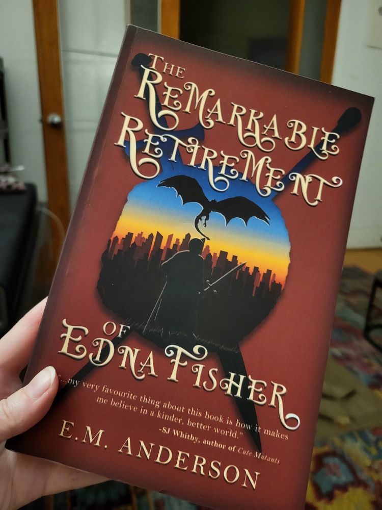 A hand on the left holds the paperback copy of The Remarkable Retirement of Edna Fisher by E.M. Anderson in the foreground.