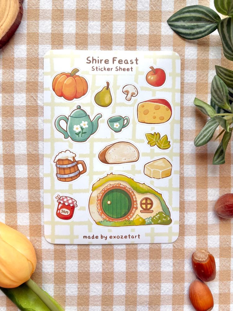 Photography of a sticker sheet called “shire feast” showing hobbit inspired food in a cute stylized way. There is a hobbit hole, beer, butter, cheese, jam, tea pot, leaves, apple, pear and mushroom.