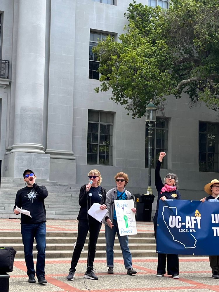 Alt text: A group of six people stand on the steps outside a large stone building, participating in a public demonstration. Three people in the center hold microphones and papers, one holds a sign reading “People with Grad Student Power,” and two others hold a large blue banner that says “UC-AFT Local 1474.” One person raises their fist in the air.