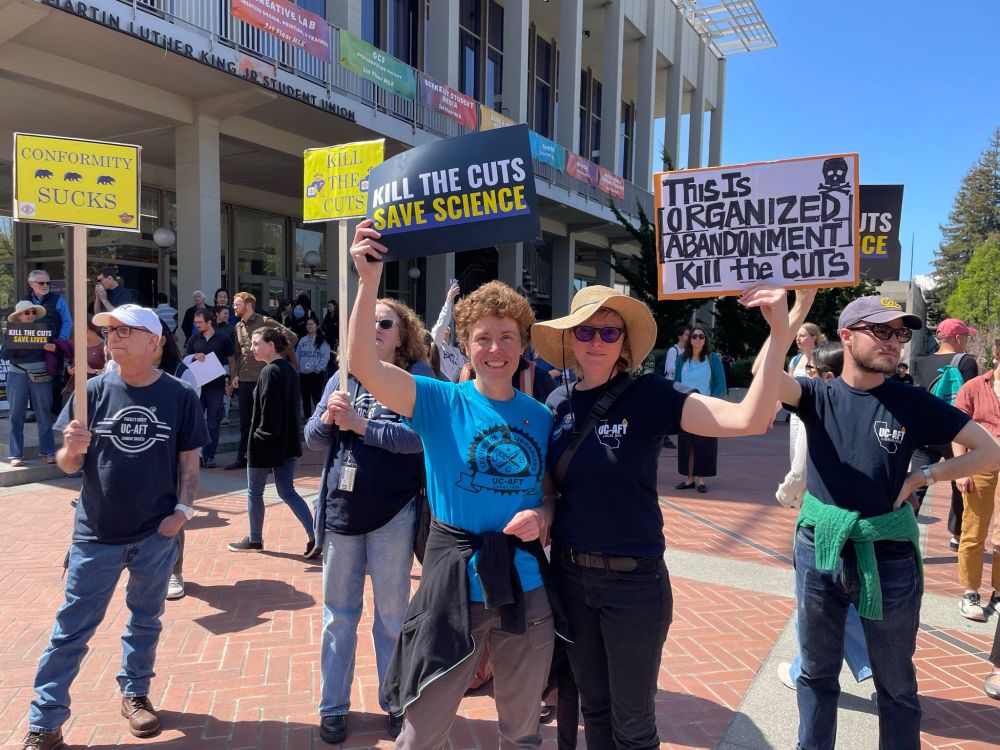 protestors in blue t-shirts stand in the sun at a rally and hold signs such as "Kill the Cuts: Save Science"