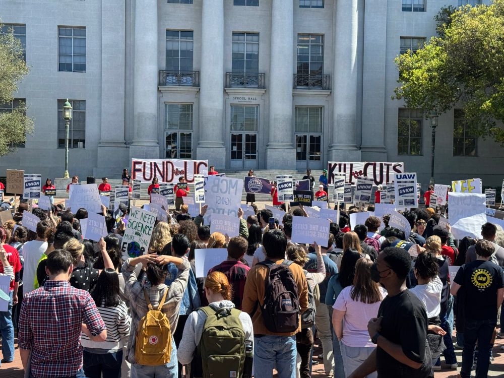 a large demonstration in front of a university building. protestors hold up signs and listen to speakers.