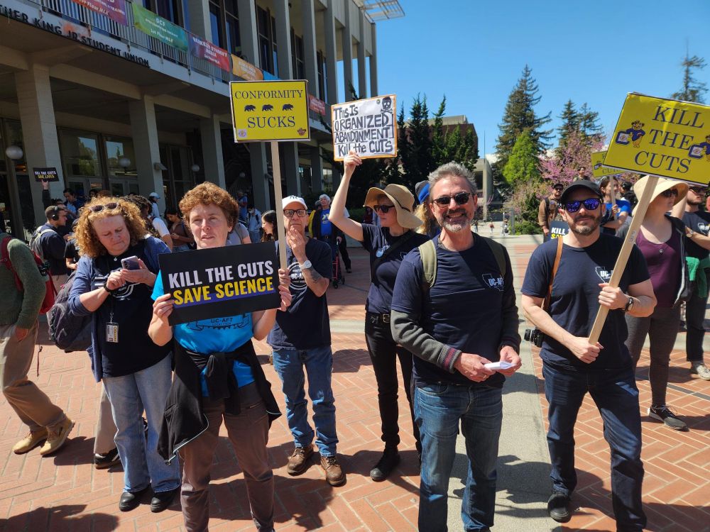 protestors in blue t-shirts stand in the sun at a rally and hold signs such as "Kill the Cuts: Save Science"