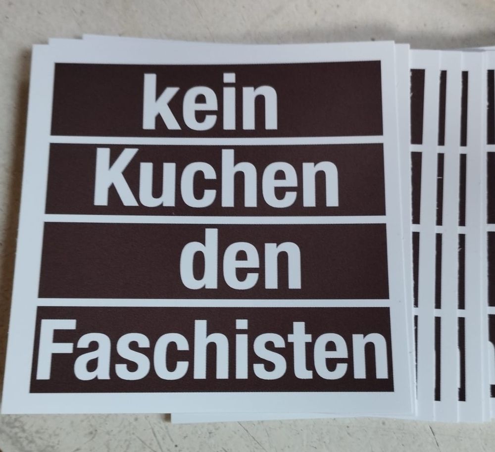 Auf einer antikweißen Oberfläche liegen mehrere Sticker . Sie sind braun mit weißem Rand. In weißer Schrift steht darauf geschrieben: kein Kuchen den Faschisten 