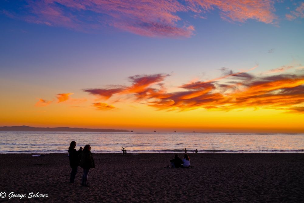View of the sunset looking across a dark, sandy beach.  The sky is orange near the horizon, shading into blue near the top of the image.  The clouds are lit with bright orange from underneath.  There are a couple of people in the foreground, seen in silhouette, taking photos of the sunset with their phones.