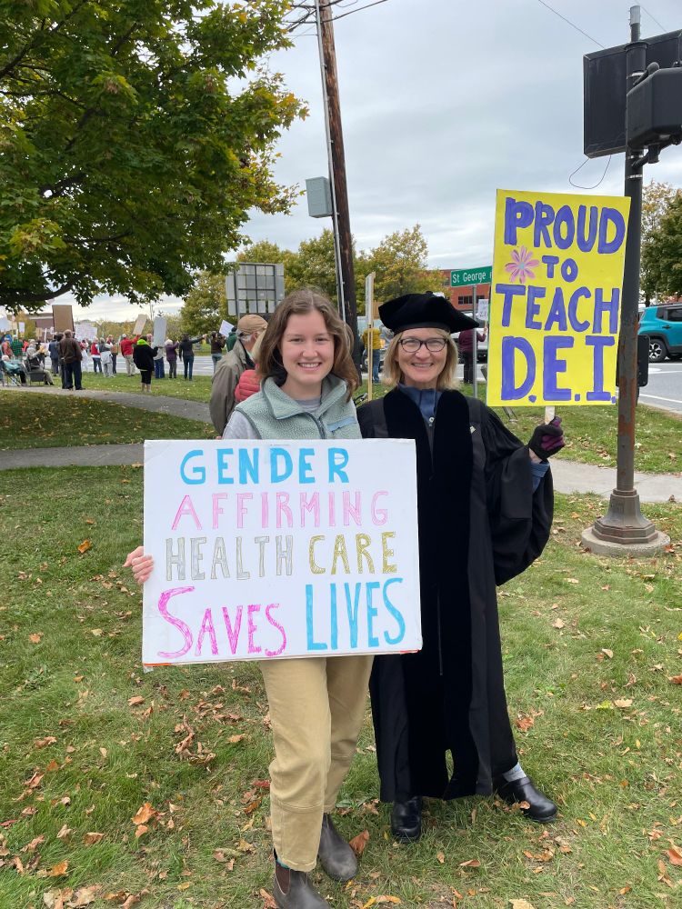 Two women at a protest, the younger one holds a sign saying "Gender affirming health care saves lives". The other woman, dressed in academic regalia, has a sign saying "Proud to teach DEI".