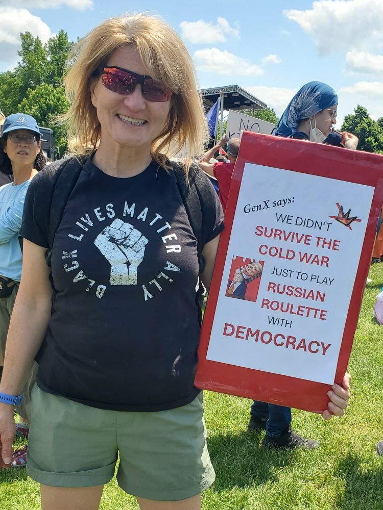 Middle aged white gal in BLM ally t-shirt holding a sign saying "GenX says: we didn't survive the Cold War just to play Russian roulette with democracy"