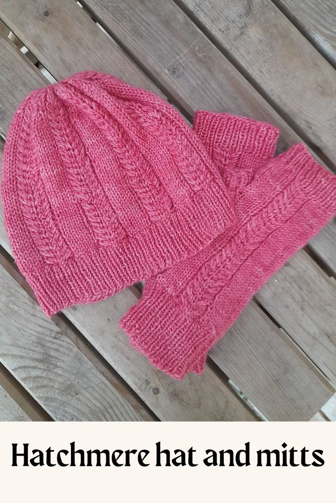 Hatchmere hat and mitts are cable knit in pink yarn. here they are resting on a table. 