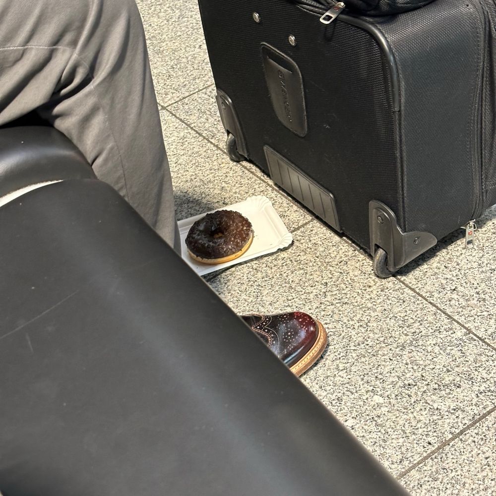 Ein Donut mit Schokolade und Schokosträuseln auf einem Pappteller auf dem Boden daneben ragt eine braune Lederschuhspitze unter einem Sitz hervor. Donut & Schuhspitze sind sich zum Verwechseln ähnlich.