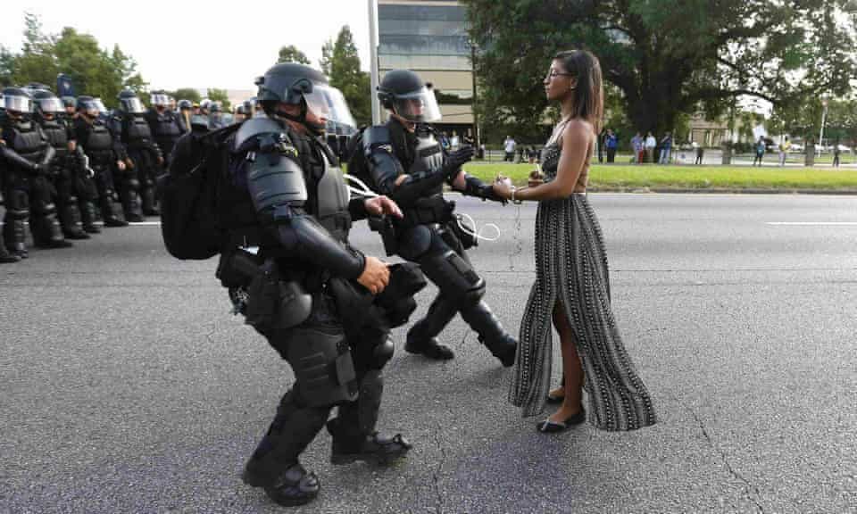 Iesha L Evans standing up to armored police in Baton Rougue. She is an elegant Black woman in a long flowing dress. A police officer is grabbing her arm. There is a row of officers before the 2 armored thugs.