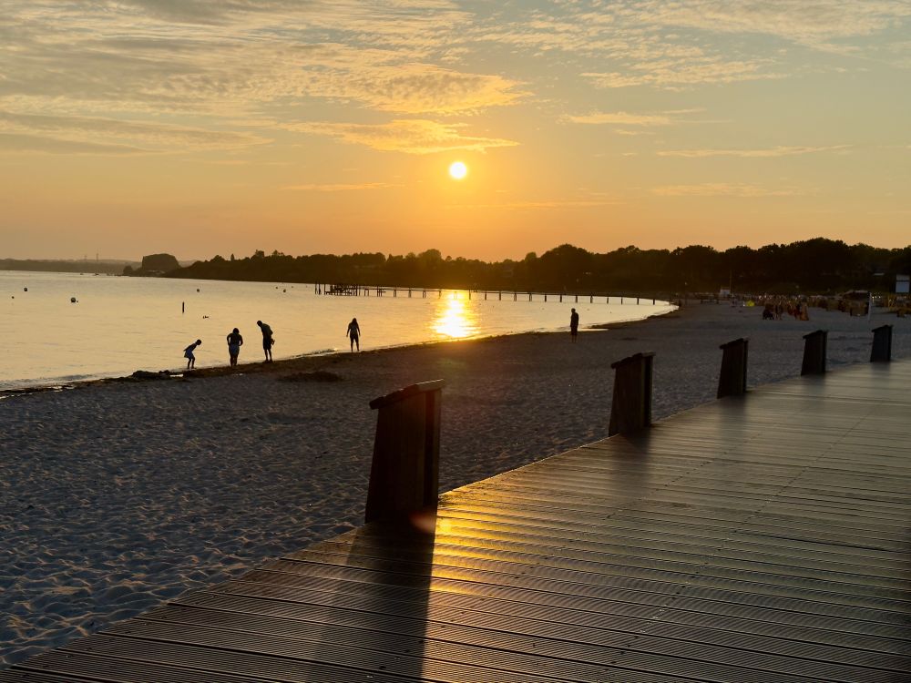 Ein Steg mit dicken niedrigen Pfosten (auf der dem Strand zugewandten Seite) die ihre Schatten auf seine glänzende Oberfläche werfen führt von der linken unteren Ecke des Bildes weiter zur Mitte des rechten Bildrandes. Die Sonne steht tief am Horizont und spiegelt sich golden im ruhigen Meer. Mehrere Personen stehen als Silhouetten am Ufer und im seichten Wasser der Bucht, von der im Hintergrund eine mit großen Bäumen bewachsene Küstenlinie zu sehen ist. Davor erstreckt sich in längerer Holzsteg vom Ufer aus nach links ins Wasser.​​​​​​​​​​​​​​​​ Der Himmel zeigt warme gelb-orange Farbtöne mit Wolken.