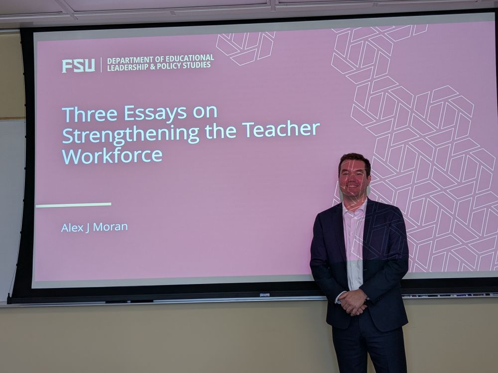 Alex smiling in front of his dissertation intro slide, which says, "Three Essays on Strengthening the Teacher Workforce"