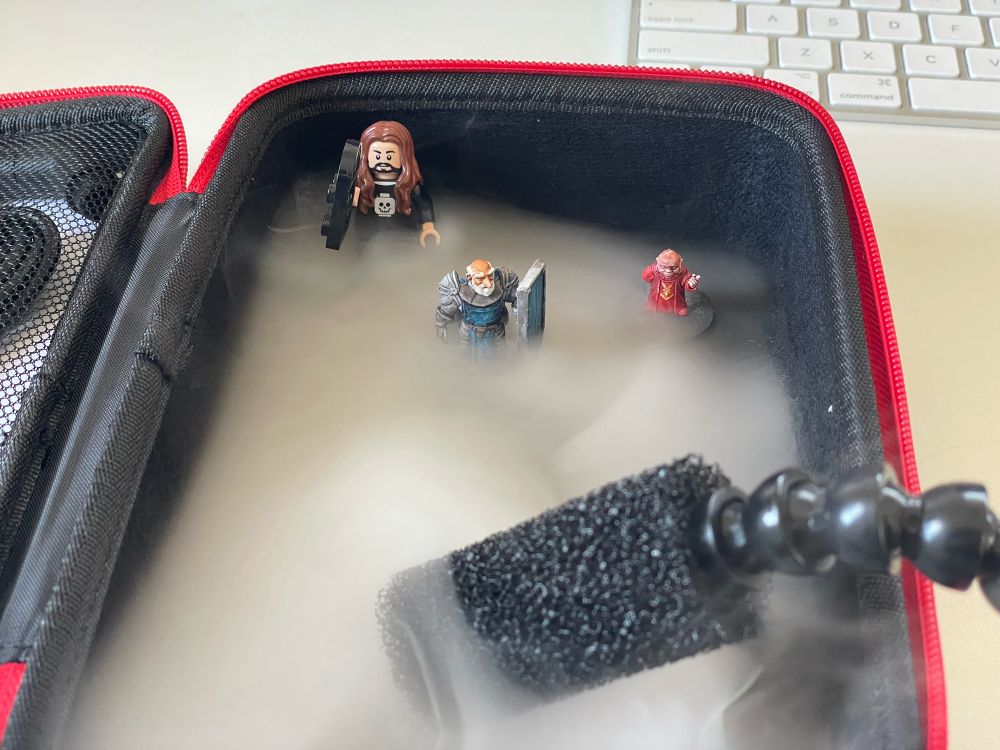 A fog machine pouring dry ice style smoke into a carry case, with two miniature figures and a Lego figure stood in the smoke.