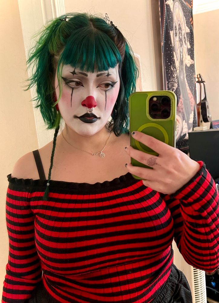 Mami Treez in an off the shoulder, black and red stripped long sleeve. Their face is in clown with a red nose, rosy cheeks, black lips and stripes and black pointy shapes. They have cute short green hair with lil long braids 