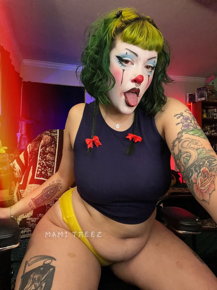 photo of mami treez in clown makeup, a blue tank top and yellow underwear. their clown makeup has a bright red nose, rosy cheeks and blue eyeshadow. their hair is green with small red bows on their braid. treez in on their knees on their official chair, showing their thick thighs, soft belly heavy tots and tattooed skin. she’s both alluring and silly all at once and you can help but to smile in a horny way about it 