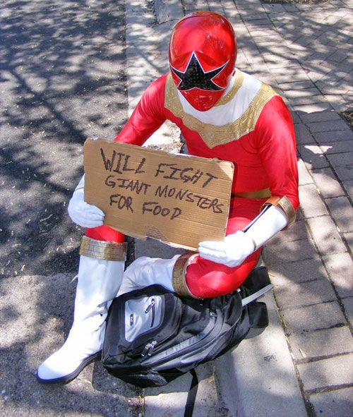 OhRed also known as the Red Zeo Ranger holding a cardboard sign saying "WILL FIGHT GIANT MONSTERS FOR FOOD" in all capitals while sitting on the street curb with a backback under their legs. 
