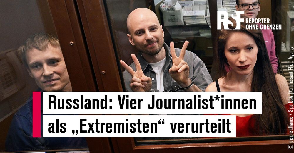 Zu sehen sind zu sehen drei Männer und eine Frau hinter einer Glasscheibe. Die Frau und ein Mann schauen traurig. Ein Mann trägt Handschellen und zeigt mit seinen Händen Peace-Zeichen, er schaut schelmisch.