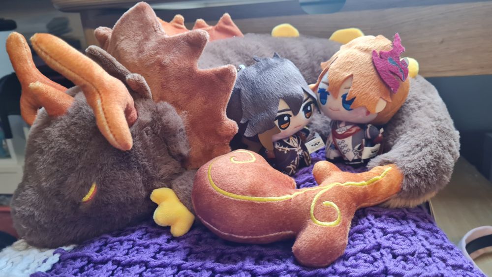 Zhongli and childe chibigurumis facing each other, resting in the coil of a rex lapis plushie