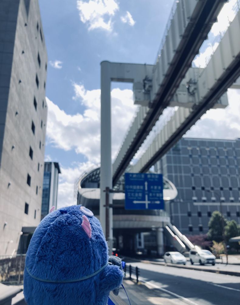 青いぬいぐるみ・モンいおが空を見上げるようにしている。背景にはモノレールの高架があり、近代的な建物が並ぶ。青い案内標識には「中央北口」「中央南口」「文化会館」「市役所」などの文字が書かれている。