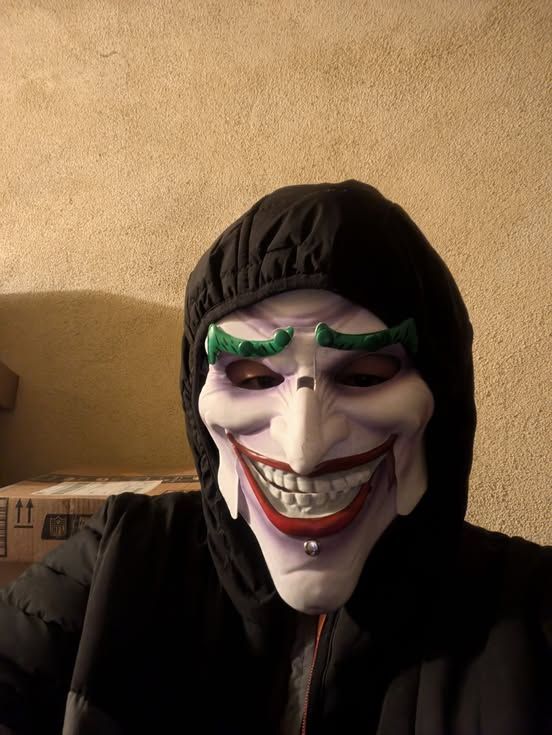 Man wearing a Halloween Mask of the Joker with a hoodie in front of a stucco wall.