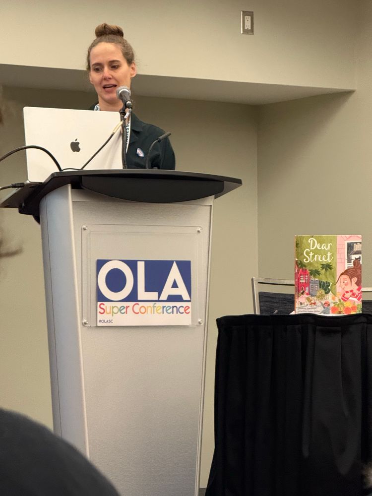 Lindsey Zier-Vogel speaking at a podium with her book “Dear  Street”