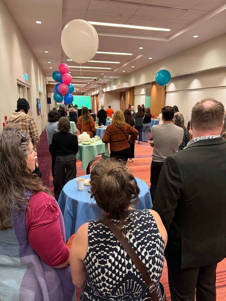 People standing around table listening to a speaker at the front of the room. Event: Tinlids Forest of Reading breakfast event.