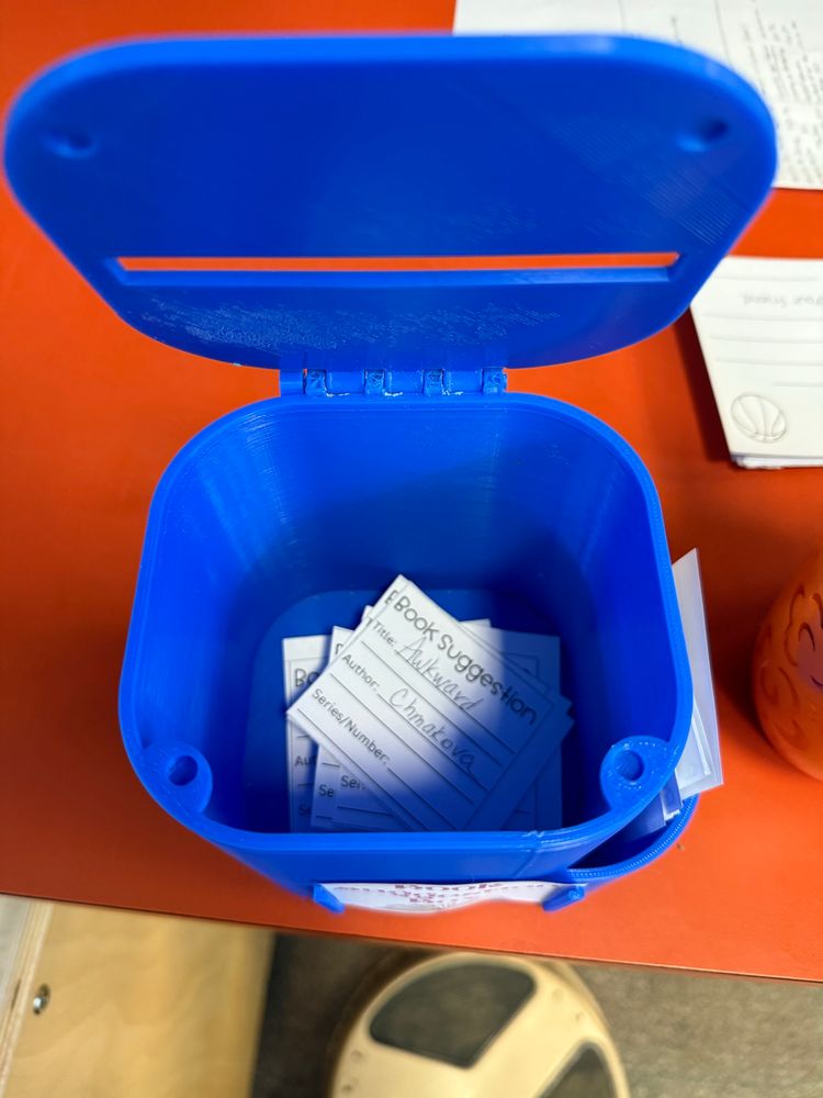 An open blue 3D printed box with small papers inside. The top paper is requesting purchasing the graphic novel “Awkward”