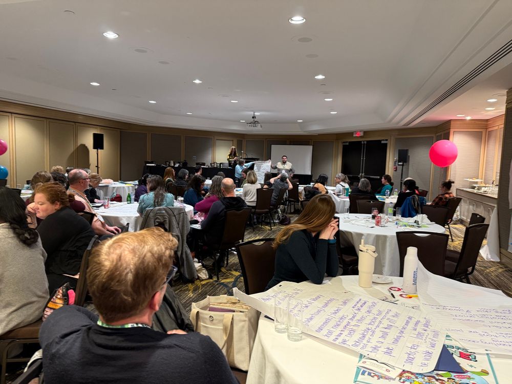 A group of people sitting around round tables with a presenter at the front. On the tables are large chart papers with brainstorming ideas written on them.