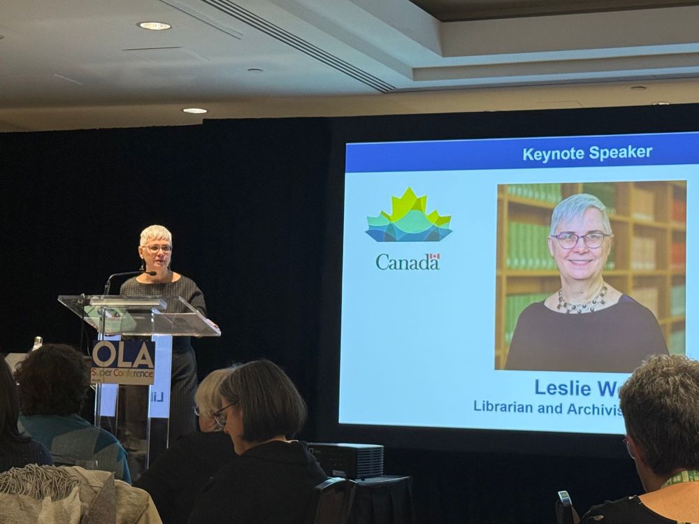 Leslie Weir, Librarian and Archivist of Canada, speaking at a lectern. There is a slide projected next to her with a photo of herself and title