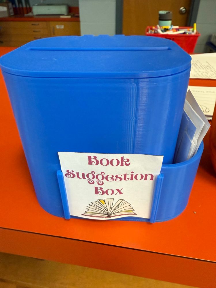 A blue 3D printed box with a sign on the front saying “Book Suggestion Box”. There is a a lot at the top for piece of paper to be inserted 