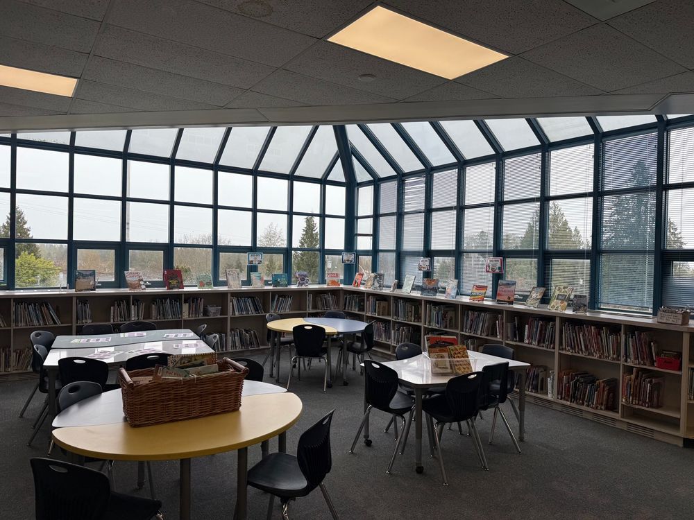 An elementary library space with tables and chairs and large solarium type windows