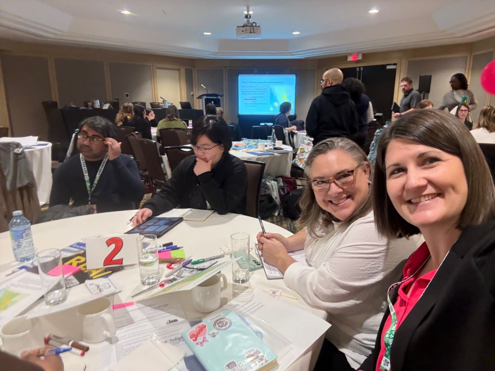 Shaunna MacDonald and Cathy Fowler taking a photo together at a table with others. The are many tables in the background also with conversations happening