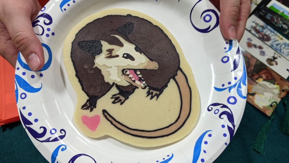 A pancake laying flat on a paper plate. The pancake is designed to look like a small screaming opossum.