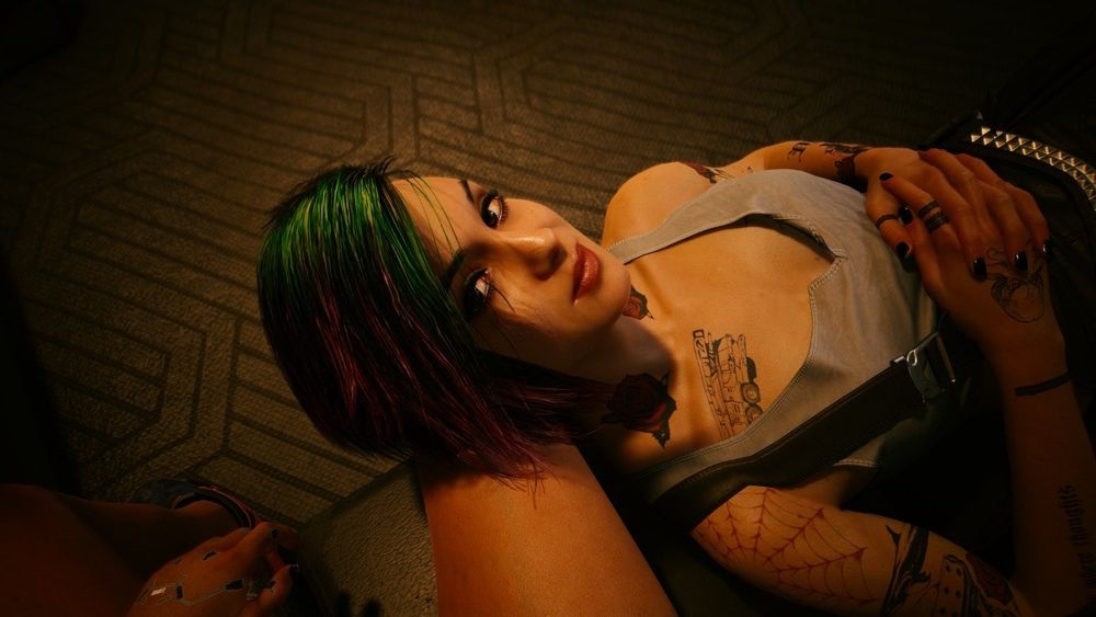 Judy, personagem de cyberpunk 2077. Ela está deitada na perna da namorada, no sofá. Ela tem pele branca, cabelo verde em cima e rosa nas pontas, caindo para um lado do rosto. Ela tem tatuagens, usa um top cinza e um macacão preto e está olhando para a foto com as mãos apoiadas no próprio peito.