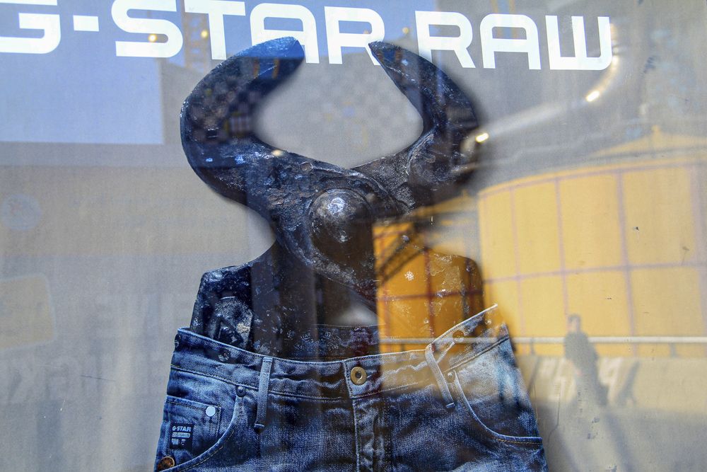 Ein Schaufenster, in dem Jeans in einer überdimensionalen Beißzange stecken. Darüber das Logo von G-Star Raw. In der Auslage spiegelt sich ein gelber Öltank der Müllverbrennungsanlage Spittelau und Personen, die davor gehen.