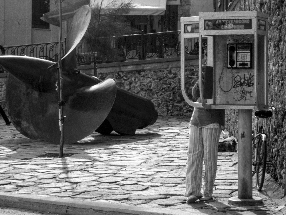 Schwarzweiß Bild. An einer offenen Telefonzelle mit zwei Telefonen telefoniert eine Frau, ihr Gesicht ist fast vollständig von der Abtrennung zwischen den beiden Telefonen verdeckt.
Sie steht an einem mit unregelmäßigen Steinen gepflasterten kleinen Platz, neben ihr eine riesiges schwarzes Teil, das für Laien auschaut wie eine Turbine.