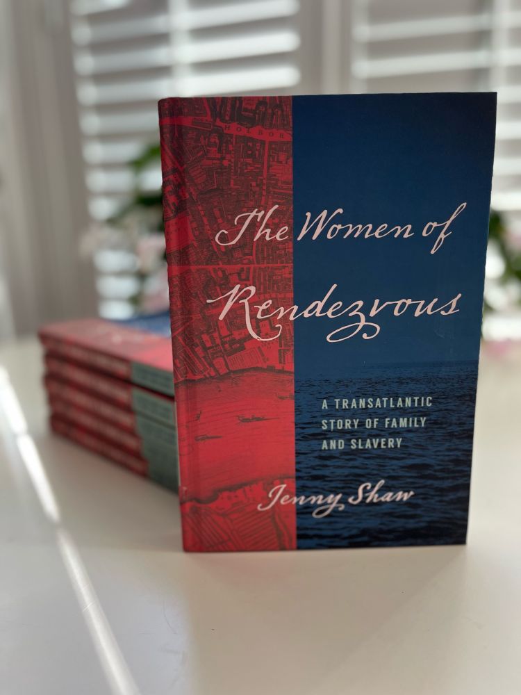Red and blue cover book (The Women of Rendezvous: A Transatlantic Story of Family and Slavery) standing upright on a white table with a stack of five books behind (not in focus)