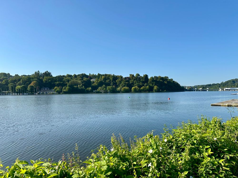 Bild des Sees mit der Sonne im Rücken, der Himmel ist strahlend blau, man sieht die Uferbewachsung und ein paar Anlegestege. Im Hintergrund Wald und in der Ferne ein Wehr.