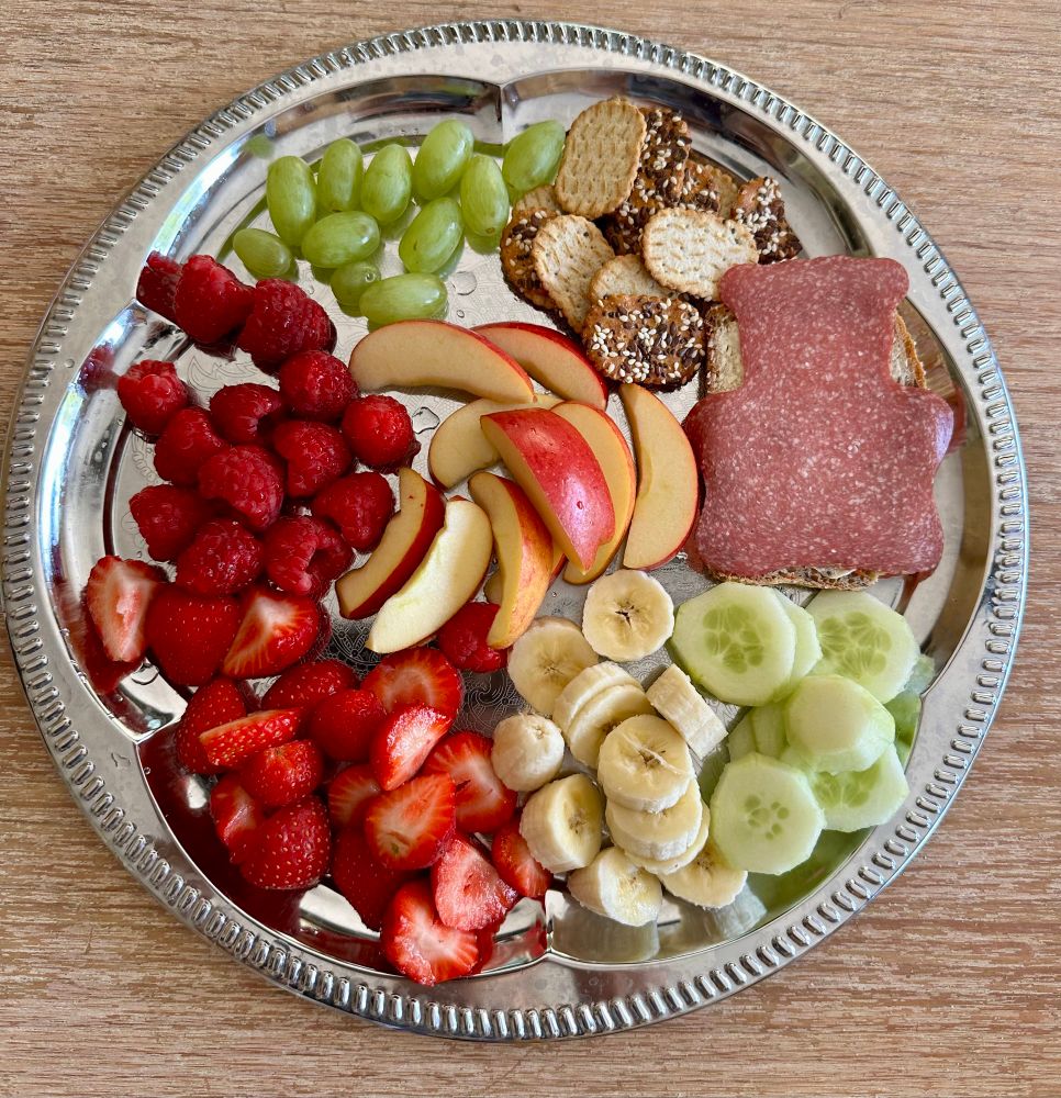 Eine silberne Servierplatte voll mit Erdbeeren, Himbeeren, Bananenstücken, Apfelschnitzen, Gurken, Trauben, Crackern und Brot.