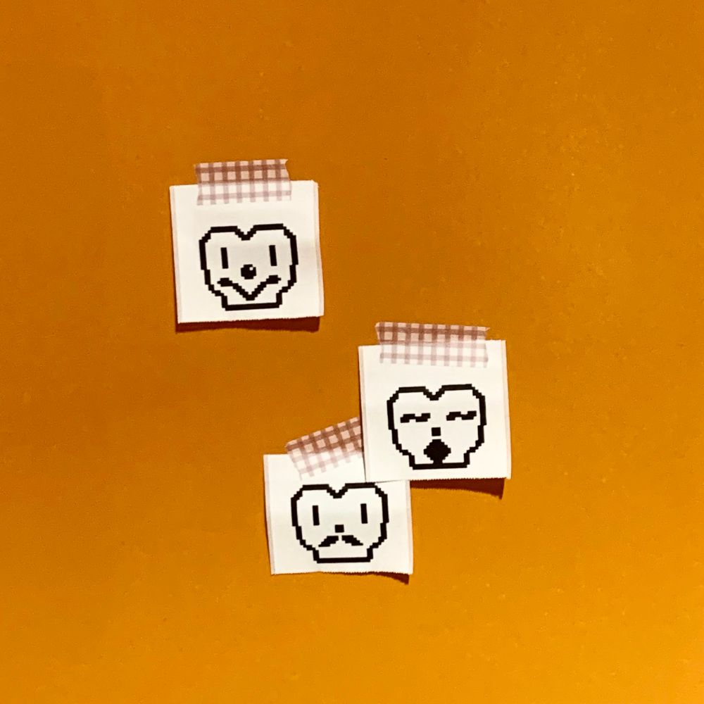 オレンジ色の壁に、印刷されたシールがマスキングテープで貼ってある。
山がふたつあるようにごはんを盛ったお茶碗のようなシルエットのキャラクターがシールに印刷されており、道化師のような顔のもの(一番上)、目が〜の記号のようになっているもの(真ん中)、ヒゲが生えているもの(一番下)がある。