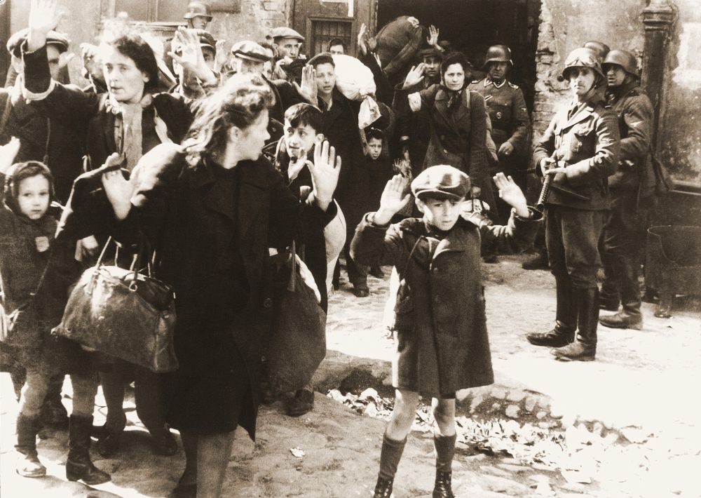 Young Polish boy standing with his arms raised in the Warsaw ghetto during a selection, surrounded by smiling Nazi soldiers with machine guns.