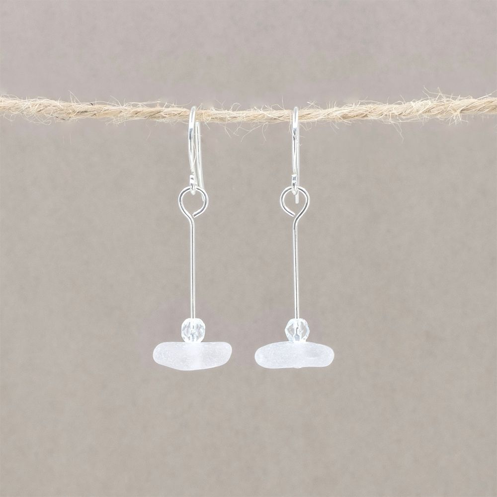 A pair of earrings where a small piece of white sea glass hangs flat from a straight silver wire. On top of each piece of glass is a crystal clear glass bead with facets that sparkle in the light.
