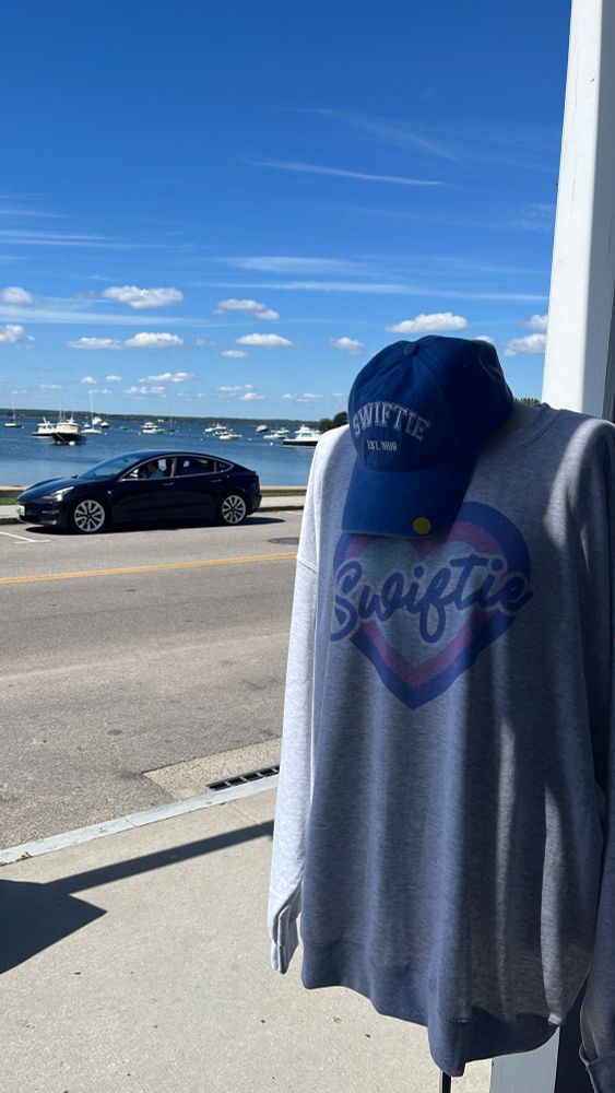 A gray sweatshirt that says Swiftie with a colorful heart and a blue baseball cap that says Swiftie est 1989 hanging outside a shop looking out on the water. Watch Hill, RI