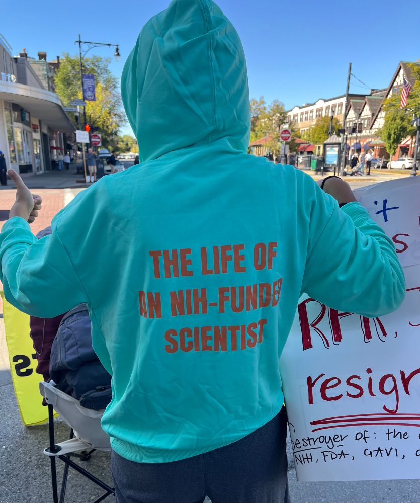 I’m wearing a mint green hoodie that says THE LIFE OF AN NIH-FUNDED SCIENTIST in orange on the back 

I’m at a protest and giving a thumbs up 