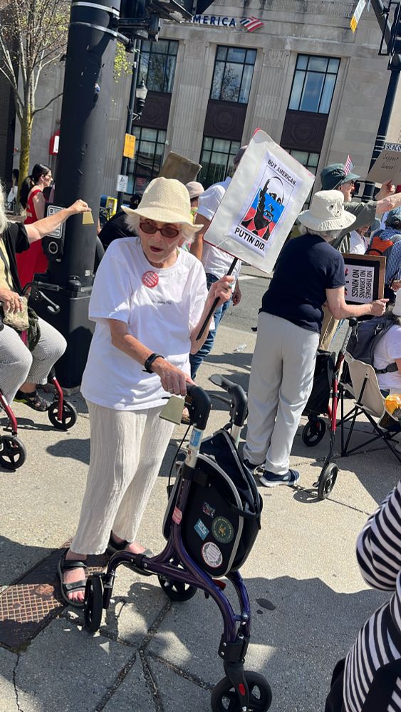 An elderly woman in white shirt, beige pants, and red lipstick with a folding walker holding up a sign that says BUY AMERICAN PUTIN DID 