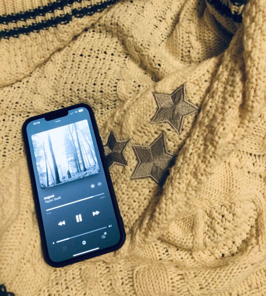 A phone showing the song “august” by taylor swift playing, laying on a white cardigan with black accents and silver elbow patches