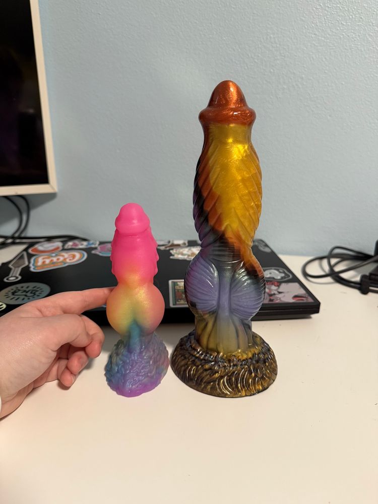A 5ish inch pastel rainbow dildo sits on a desk next to a 10ish inch gold and bronze dildo as a size comparison